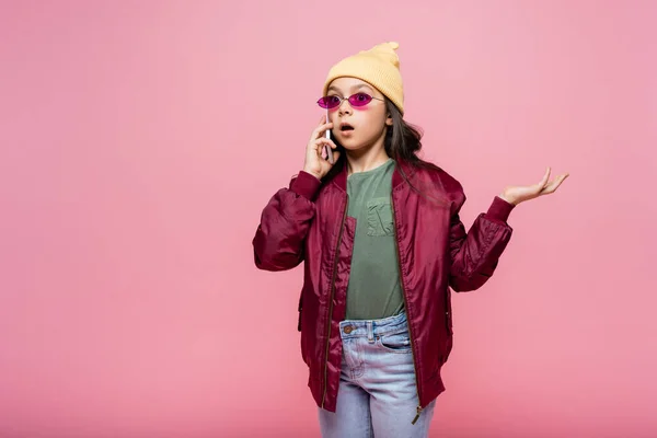 Choqué préadolescente en tenue élégante et lunettes de soleil parler sur smartphone tout en geste isolé sur rose — Photo de stock