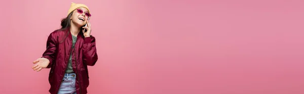 Chica preadolescente feliz en traje elegante y gafas de sol hablando en el teléfono inteligente aislado en rosa, pancarta - foto de stock