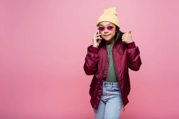 Joyful preteen girl in stylish outfit and sunglasses talking on smartphone and showing thumb up isolated on pink — Stock Photo
