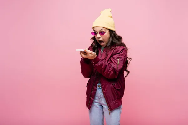 Angry preteen girl in stylish outfit and sunglasses looking at smartphone and screaming isolated on pink — Stock Photo