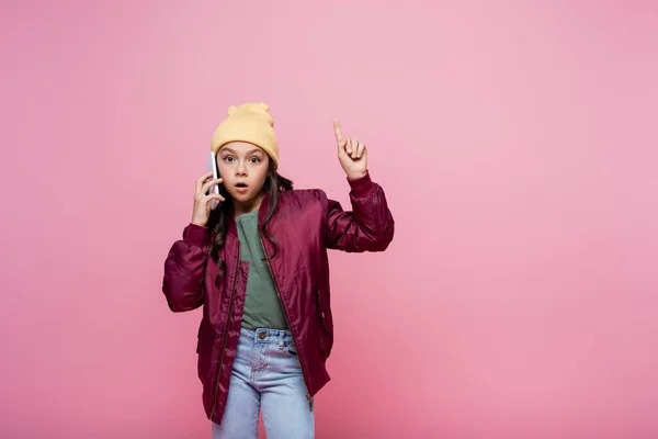 Preteen girl in stylish outfit talking on cellphone and pointing with finger isolated on pink — Stock Photo