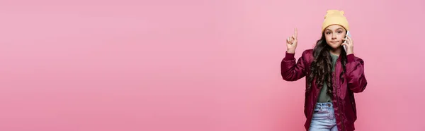 Preteen Mädchen in stylischem Outfit spricht auf Handy und zeigt mit dem Finger isoliert auf rosa, Banner — Stockfoto
