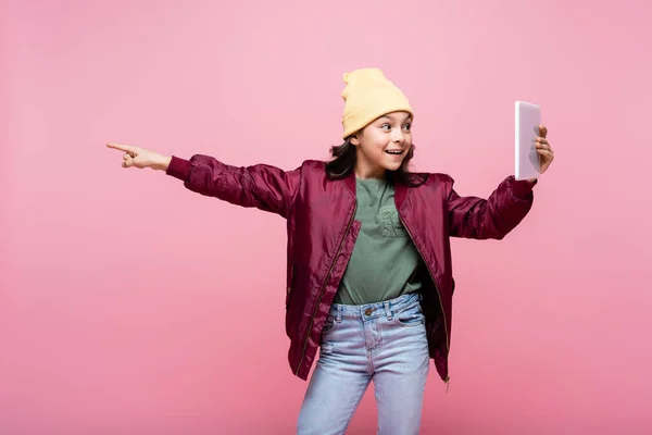 Feliz niña preadolescente en traje de moda sosteniendo tableta digital y apuntando con el dedo aislado en rosa - foto de stock