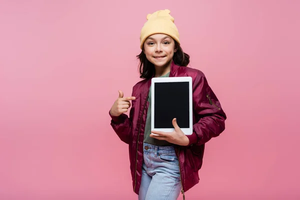 Счастливая девочка-подросток в модном наряде, указывая пальцем на цифровой планшет с чистым экраном, изолированным на розовый — стоковое фото