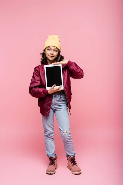 Повна довжина усміхненої дівчини в зимовому вбранні, що тримає цифровий планшет з порожнім екраном на рожевому — стокове фото