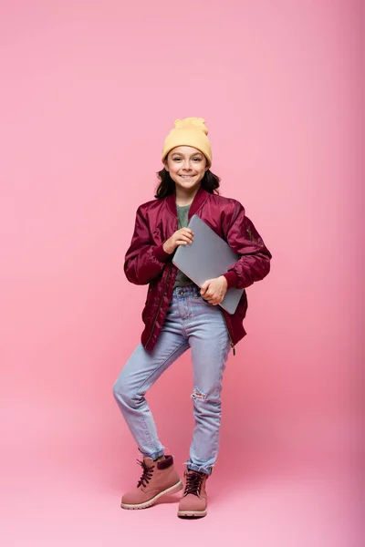 Full length of stylish preteen girl in winter outfit posing with laptop and smiling on pink — Stock Photo