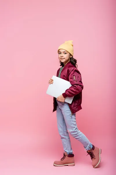 Comprimento total da menina pré-adolescente elegante em roupa de inverno posando com laptop em rosa — Fotografia de Stock