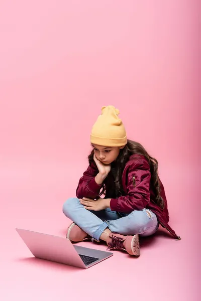 Longitud completa de la chica preadolescente con estilo en traje de invierno sentado y mirando a la computadora portátil en rosa - foto de stock