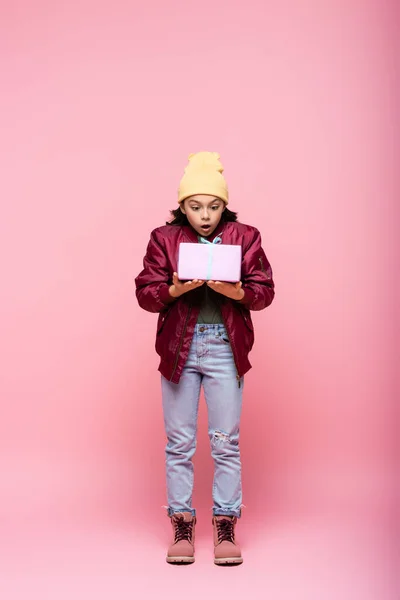 Comprimento total de menina pré-adolescente surpresa em roupa de inverno olhando embrulhado presente em rosa — Fotografia de Stock