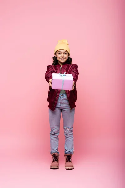 Longitud completa de la niña preadolescente feliz en traje de invierno sosteniendo regalo envuelto en rosa - foto de stock