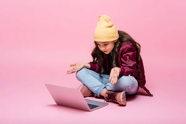 Élégant preteen fille en tenue d'hiver assis et regardant ordinateur portable et montrant geste haussant les épaules sur rose — Photo de stock