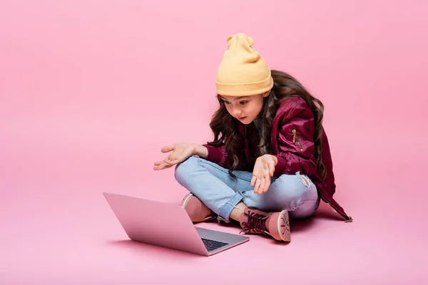 Stylish preteen girl in winter outfit sitting and looking at laptop and showing shrug gesture on pink — Stock Photo