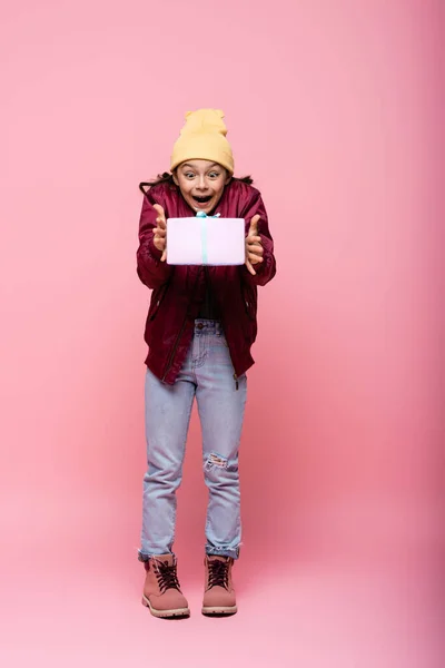 Повна довжина здивованої дівчини в зимовому вбранні, дивлячись на загорнутий подарунок на рожевому — стокове фото