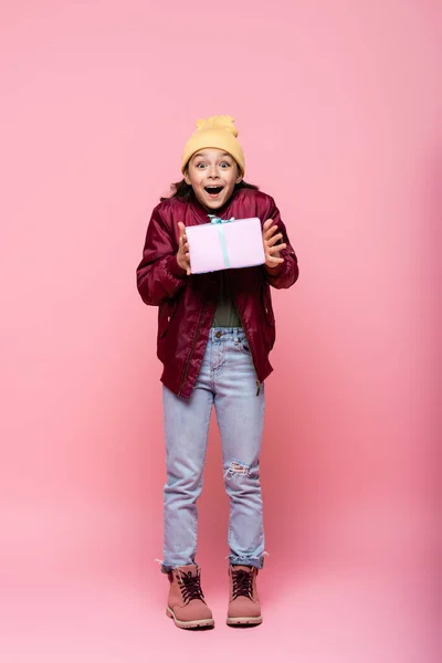 Full length of amazed preteen girl in winter outfit holding wrapped present on pink — Stock Photo