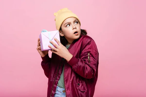 Menina pré-adolescente curioso na roupa da moda segurando presente envolto isolado em rosa — Fotografia de Stock