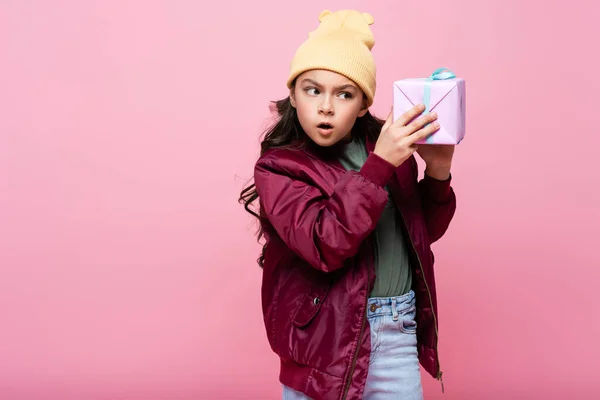 Curious preteen kid in trendy outfit shaking wrapped present isolated on pink — Stock Photo