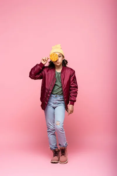 Full length of stylish preteen girl in winter outfit covering eye with lollipop on pink — Stock Photo