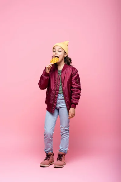 Full length of preteen girl in stylish outfit eating lollipop on pink — Stock Photo