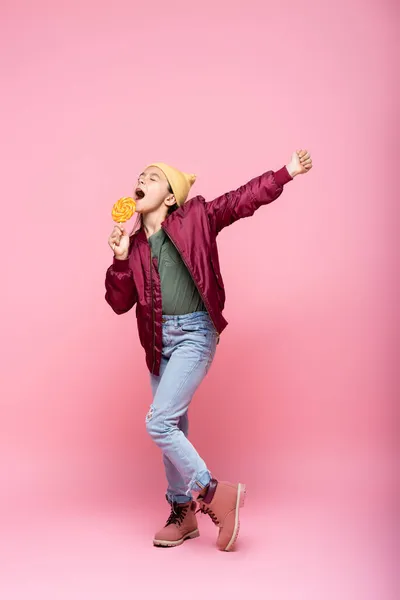 Full length of stylish preteen girl in winter outfit holding lollipop and singing on pink — Stock Photo