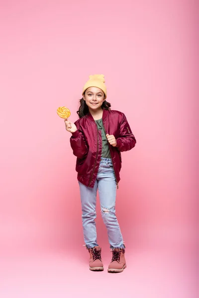 Longitud completa de la niña preadolescente feliz en traje de invierno celebración de piruleta en rosa - foto de stock