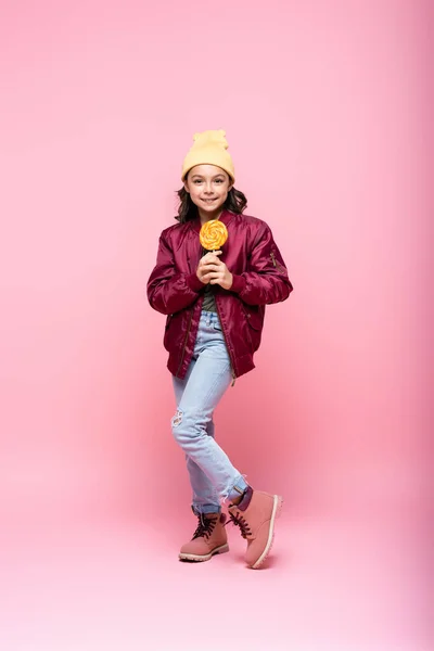 Full length of cheerful preteen girl in winter outfit holding lollipop on pink — Stock Photo
