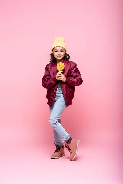 Longitud completa de alegre chica preadolescente en traje de invierno celebración de piruleta en rosa — Stock Photo