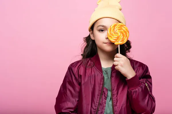 Stylish preteen girl in winter outfit holding lollipop and covering eye isolated on pink — Stock Photo