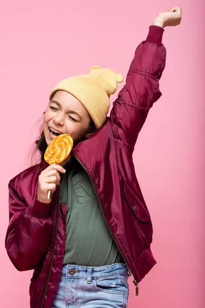 Cheerful preteen girl in winter outfit holding lollipop and smiling isolated on pink — Stock Photo