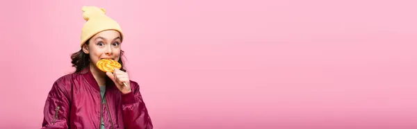 Стильна дівчина в зимовому вбранні кусає льодяник ізольовано на рожевому, банер — стокове фото