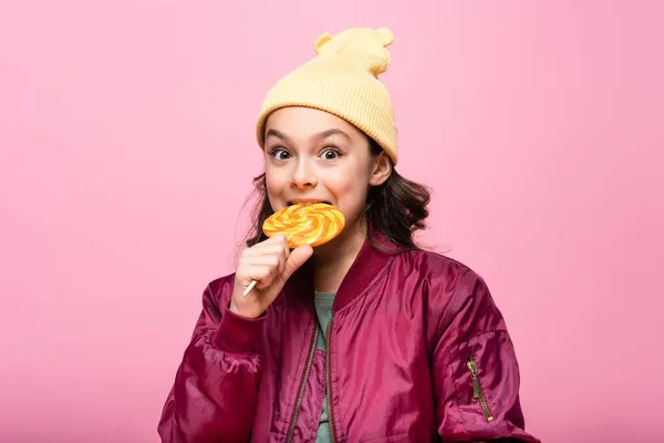 Trendy preteen girl in winter outfit biting lollipop isolated on pink — Stock Photo