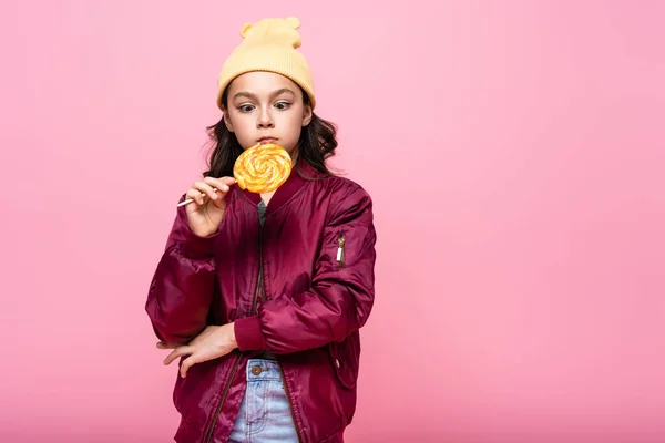 Stylish preteen girl in winter outfit looking at lollipop isolated on pink — Stock Photo