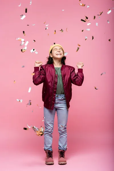 Pleine longueur de heureux preteen fille en tenue élégante souriant près de tomber confettis sur rose — Photo de stock