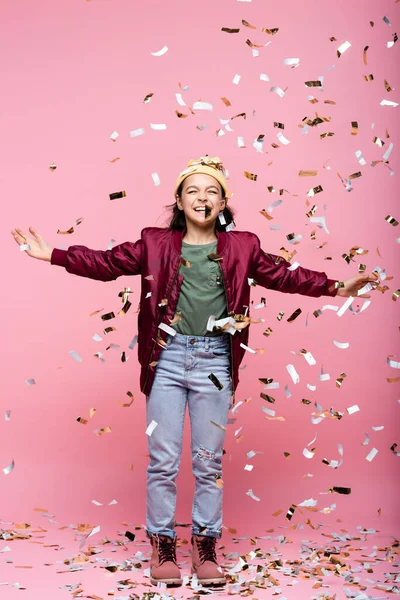 Pleine longueur de gai preteen fille en tenue élégante souriant près de tomber confettis sur rose — Photo de stock