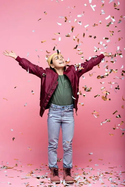 Full length of joyful preteen girl in stylish outfit smiling near falling confetti on pink — Stock Photo