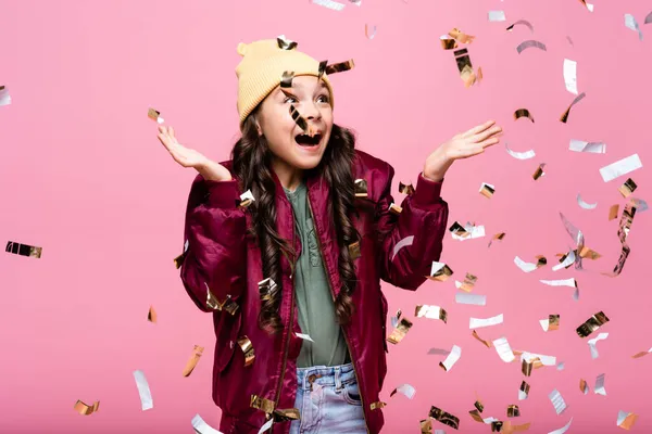 Amazed preteen girl in stylish outfit smiling near falling confetti on pink — Stock Photo