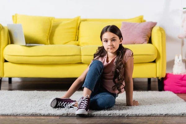 Preteen menina em jeans olhando para a câmera e sentado no tapete na sala de estar — Fotografia de Stock