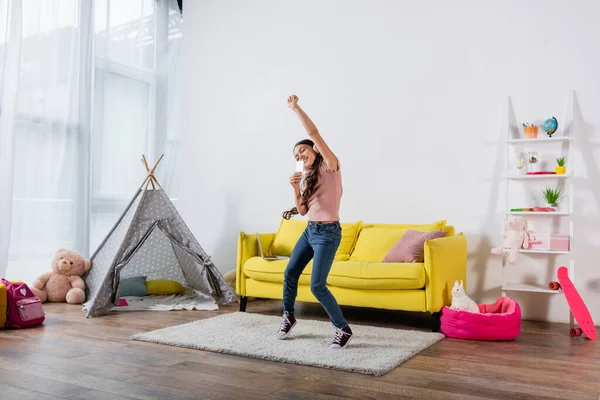 Glückliches Mädchen mit drahtlosen Kopfhörern, das das Smartphone hält, während es im modernen Wohnzimmer tanzt — Stockfoto