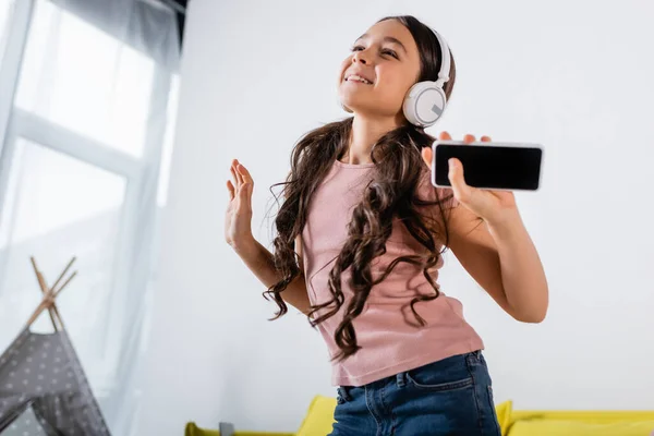 Happy preteen girl in wireless headphones holding smartphone while dancing at home — Stock Photo