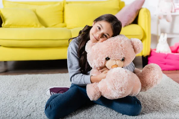 Alegre preteen menina abraçando brinquedo macio enquanto sentado no tapete em casa — Fotografia de Stock