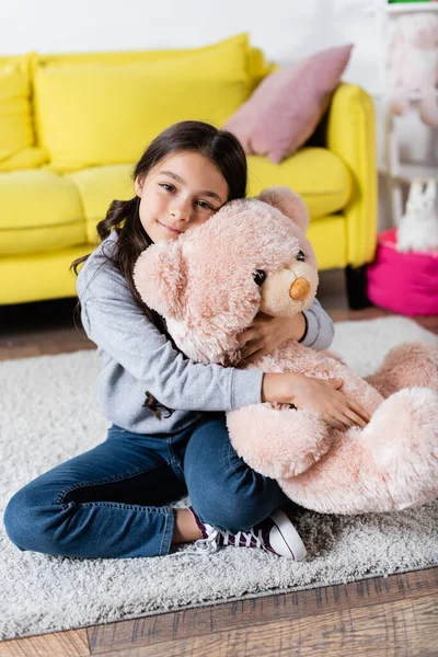 Complacido niña preadolescente abrazando juguete suave mientras está sentado en la alfombra en casa - foto de stock