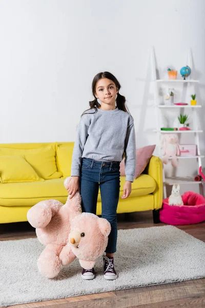 Niña preadolescente sosteniendo juguete suave mientras está de pie en la alfombra en casa - foto de stock