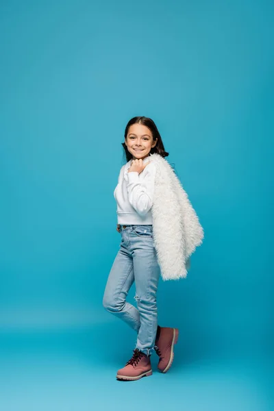 Pleine longueur de fille preteen heureux en sweat à capuche et jeans tenant veste blanche en fausse fourrure sur bleu — Photo de stock