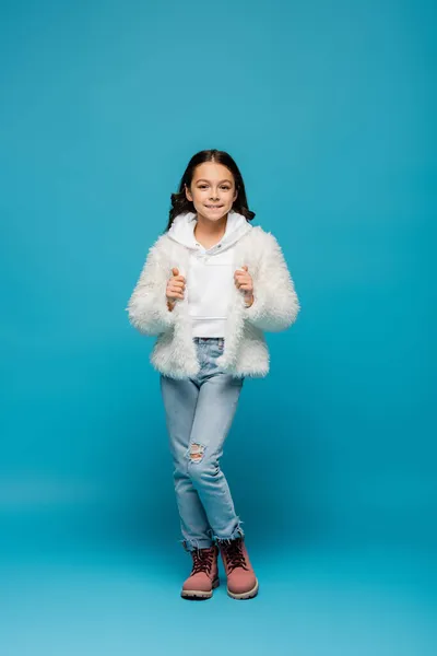 Voller Länge positiver Frühchen in Kunstpelzjacke und Winterstiefeln, die auf blau posieren — Stockfoto