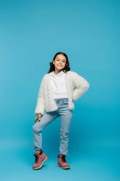 Full length of positive preteen kid in faux fur jacket and winter boots posing with hand on hip on blue — Stock Photo