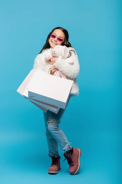 Pleine longueur de jeune fille heureuse en lunettes de soleil et veste en fausse fourrure étreignant lapin doux et tenant des sacs à provisions sur bleu — Photo de stock