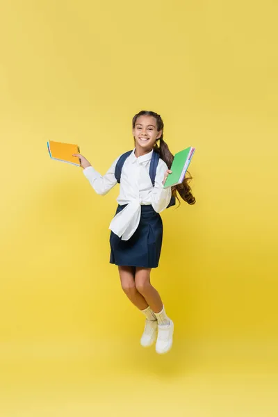 Smiling schoolgirl with notebooks jumping and looking at camera on yellow — Stock Photo