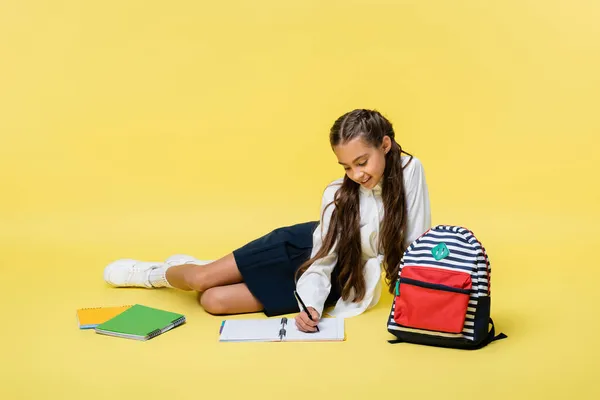 Criança sorridente escrevendo no caderno perto da mochila no fundo amarelo — Fotografia de Stock