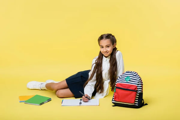 Schoolgirl writing on notebook and smiling at camera near backpack on yellow background — Stock Photo