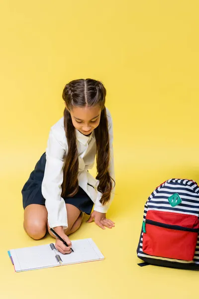 Lächelndes Schulkind schreibt auf Notizbuch neben Rucksack auf gelbem Hintergrund — Stockfoto