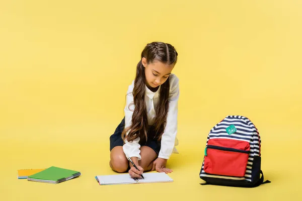 Schoolgirl writing on notebook near backpack on yellow background — Stock Photo